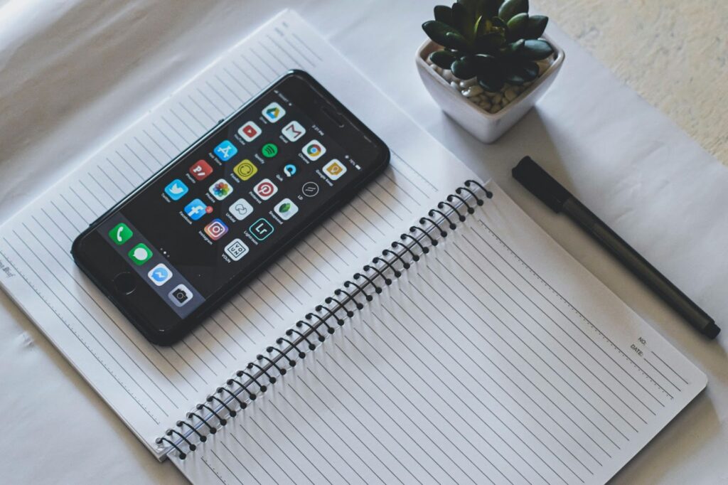 A smartphone on a table with its screen displaying various app icons, accompanied by a notebook and pen placed beside it