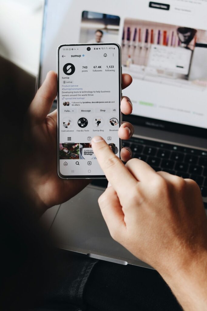 A person holding a phone and looking at an Instagram profile, with a laptop in the background