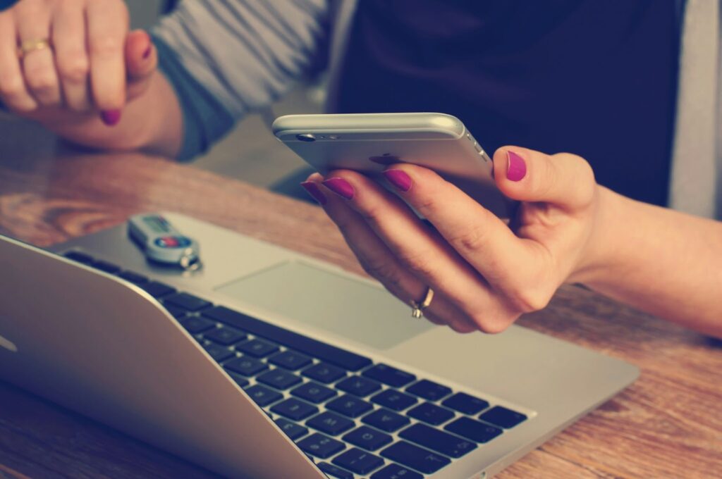 A woman holding a phone with a laptop placed on the table in front of her.